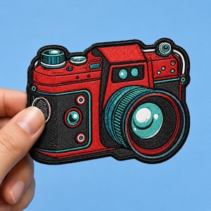 May include: An embroidered patch shaped like a vintage camera, featuring a vibrant red body with black textured side panels and teal blue accents. The design includes a prominent circular lens, various dials, and buttons, all outlined in black thread.