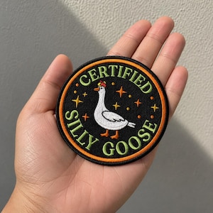 May include: A circular embroidered patch with a black background and an orange border. The center features a white goose wearing a small red hat, surrounded by orange stars. The text reads CERTIFIED SILLY GOOSE in light green capital letters.