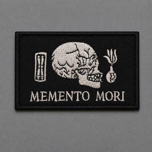 May include: Black rectangular patch with white embroidered skull, torch, and book illustrations. The phrase "MEMENTO MORI" is embroidered in white below the illustrations. The patch is likely for clothing or accessories.