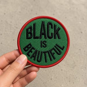 May include: A circular embroidered patch featuring a green background with a red border. The text "BLACK IS BEAUTIFUL" is stitched in black thread across the center of the patch in a bold, sans-serif font style.