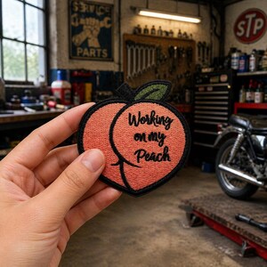 May include: Embroidered patch in the shape of a peach with the text "Working on my Peach". The patch is peach-colored with a black border and a green leaf. The patch is held in front of a garage setting.