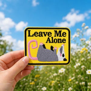 May include: A yellow patch with the text "Leave Me Alone" and a cartoon opossum. The opossum is gray with a pink tail and paws, lying on its back. The patch has a black border.
