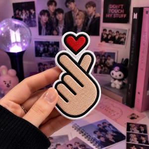 May include: An embroidered patch featuring a tan hand making a finger heart gesture with a red heart shape above the fingers. The patch has a thick white and black border, showcasing detailed stitching throughout the design of the hand and heart.