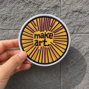 May include: Round embroidered patch with a yellow sunburst design and the words "make art" in black. The patch has a white border and is held against a gray textured surface.