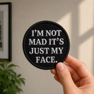 May include: A circular black embroidered patch featuring the white text I'M NOT MAD IT'S JUST MY FACE. The patch has a thick black stitched border and a textured fabric surface, displaying clear white lettering in a bold, sans-serif font style.