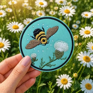 May include: Round embroidered patch featuring a bee in flight over a thistle. The patch has a teal background with a black border. The bee is black and yellow with brown wings. The thistle is white and green. Daisies are in the background.