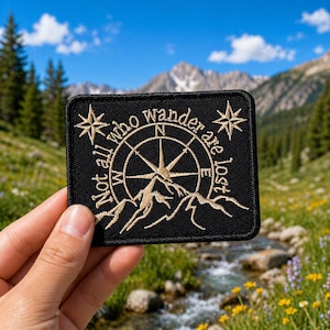 May include: Black rectangular patch with the text "Not all who wander are lost" surrounding a compass rose and mountain graphic in tan. Two starbursts flank the text. The patch is held up in front of a mountain landscape.