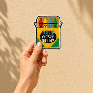May include: Embroidered patch featuring a yellow crayon box with four colorful crayons. The patch has the text "COLOR OUTSIDE THE LINES" in a playful font. The patch is outlined in white and has a green and black border.