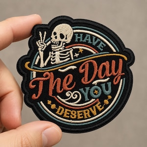 May include: A circular embroidered patch with a black background featuring a white skeleton making a peace sign. The text reads HAVE THE DAY YOU DESERVE in orange, teal, and yellow thread, accented by circular lines and small star shapes.