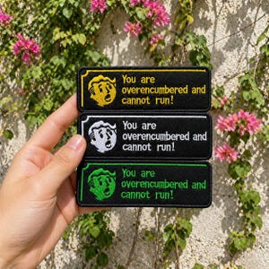 May include: Three black embroidered patches with the text "You are overencumbered and cannot run!". Each patch features a different colored border and character: yellow, white, and green. The patches are rectangular.
