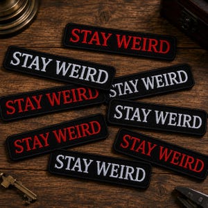 May include: A collection of rectangular black embroidered patches featuring the text STAY WEIRD in either bold red or white thread. Each patch has a clean, stitched border and is arranged in a scattered pattern across the wooden surface.