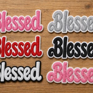 May include: Six embroidered patches with the word "Blessed" in various colors: pink, gray, red, black, white, and a lighter pink. Each patch has a white border and is displayed on a wooden surface.