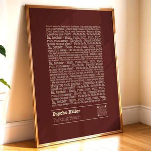 May include: A framed maroon print with the lyrics of "Psycho Killer" by Talking Heads in white. The gold-framed artwork is displayed on a light wall and wooden floor. The lyrics are vertically arranged. The print includes the band's name.
