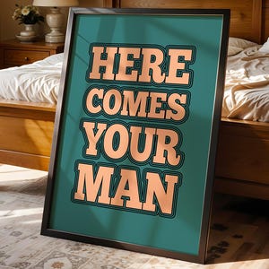 May include: A framed art print featuring the words "HERE COMES YOUR MAN" in a retro peach font with a black outline. The text is set against a teal background. The black-framed artwork is displayed in a bedroom, leaning against a bed.