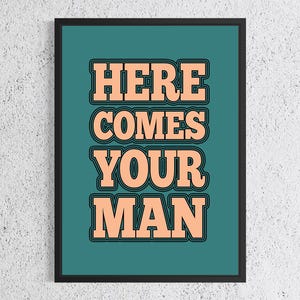 May include: A framed art print featuring the text "HERE COMES YOUR MAN" in peach-coloured, bold, retro-style lettering with black outlines, set against a teal background. The print is in a black frame.