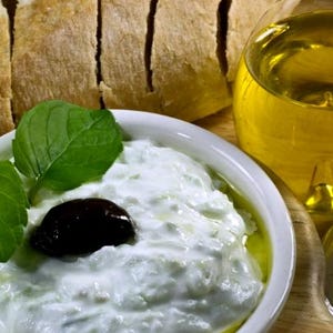 Könnte beinhalten: Ein Stillleben mit einer Schüssel Tzatziki mit einer schwarzen Olive und frischen Minzblättern, geschnittenem Brot, einem Glas Olivenöl und einem Salzstreuer. Das Essen ist auf einem Holztablett angerichtet.