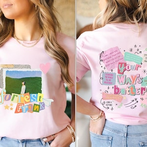 May include: A light pink t-shirt featuring a colorful graphic on the front with the words "forrest frank" and a photo of a person in a field. The back of the shirt has the words "Your Way's Better" with additional text and illustrations.