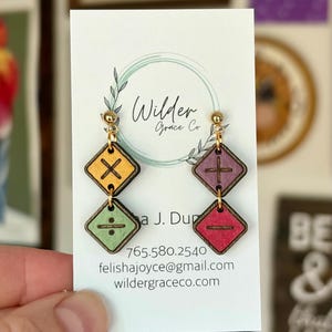 May include: Two pairs of dangling earrings featuring square wooden charms with math symbols. The left earring has a yellow multiplication sign and a green division sign. The right earring has a purple plus sign and a red minus sign. Gold hardware.