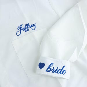Puede incluir: Una camisa blanca de botones con bordado azul. El nombre Jeffrey está bordado sobre el bolsillo del pecho, y la palabra bride junto a un símbolo de corazón está bordada en el puño de la manga con una tipografía cursiva azul.