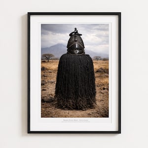 Könnte beinhalten: Ein gerahmter Fotodruck einer schwarzen Bundu (Sowei) Maske aus Sierra Leone. Die Maske hat ein glattes, dunkles, geschnitztes Kopfteil mit einer spitzen Oberseite und einen langen, dicken, schwarzen Bastfransenbesatz, der den Körper bedeckt.