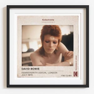 May include: Framed print featuring a vintage photograph of David Bowie. The image shows Bowie with red hair and makeup, a gold bracelet, and a white circle on his forehead. Text includes "DAVID BOWIE" and "HAMMERSMITH ODEON, LONDON JULY 1973".