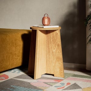 May include: A light brown wooden side table with a trapezoidal base. A small, woven basket with a handle sits on top of a book. The table is on a colorful geometric patterned rug.
