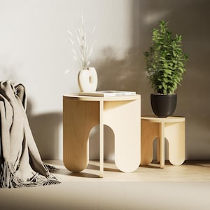 Könnte beinhalten: Zwei hellfarbene Beistelltische aus Holz mit abgerundeten Beinen und bogenförmigen Ausschnitten. Auf einem Tisch steht eine weiße Vase mit Trockenblumen und einem Buch. Der andere Tisch hat eine grüne Topfpflanze. Links ist ein grauer Stuhl mit einer Decke.