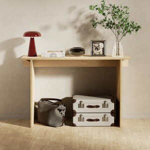 Op de afbeelding: Een lichte houten console met een rode lamp, boeken, een zwarte kom en een fotolijstje bovenop. Daaronder een grijze handtas en twee gestapelde vintage koffers. Een vaas met groen voegt een natuurlijk tintje toe.