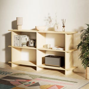 Op de afbeelding: Een lichtgekleurde houten boekenplank met meerdere vakken. De planken zijn gevuld met decoratieve items, waaronder boeken, een fotolijst, een lamp, een kom, een vaas en een luidspreker. Een patroonkleed staat op de voorgrond.