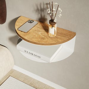 Peut inclure: Une étagère murale semi-circulaire avec une surface supérieure en bois clair et une base blanche. L'étagère supporte un smartphone, un diffuseur à roseaux en verre avec des fleurs séchées et un livre blanc portant le texte LASKASAS sur la tranche.
