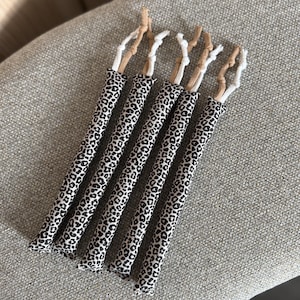 Può includere: Cinque tubi cilindrici in tessuto con una stampa leopardata bianca e nera. Ogni tubo è sormontato da due brevi passanti in tessuto annodati, di colore beige e bianco alternato, che si estendono dal bordo superiore del tessuto stampato.