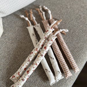 May include: A collection of long, slender fabric tubes featuring two distinct patterns: a white background with a delicate red and brown floral print, and a tan and white gingham check. Each tube is finished with a knotted brown cord at the top.