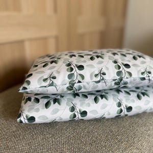 Peut inclure: Deux oreillers rectangulaires empilés l'un sur l'autre, présentant un fond blanc avec un motif répétitif de feuilles d'eucalyptus vert foncé et vert sauge ainsi que de fines tiges délicates imprimées sur la surface du tissu.
