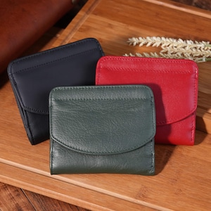 May include: Three leather wallets in navy blue, red, and green are arranged on a wooden surface. Each wallet has a flap closure. The wallets are small and compact, suitable for carrying cards and cash.