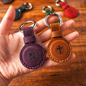 以下が含まれることがあります： 銀色のリングが付いた2つの革製キーホルダーのクローズアップ。1つは紫色のエンボス加工された「N」で、もう1つは茶色の十字架です。緑、赤、黒の他のキーホルダーが背景に見えます。
