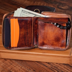 May include: Open brown leather wallet with a zip closure, filled with cash and cards. The wallet has a quote etched inside. A black beaded bracelet is visible inside the wallet.