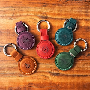 Puede incluir: Llaveros de cuero en varios colores, incluyendo burdeos, rojo, verde azulado y marrón. Cada llavero presenta un diseño circular con un llavero plateado y detalles en relieve, como iniciales, un nombre o un ancla.