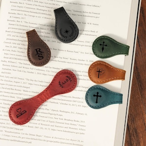 May include: Six leather magnetic bookmarks in black, brown, green, tan, teal, and red. Each bookmark features a unique laser-engraved design, including a star with laurel, initials R S, A&B, a cross, a dove with a cross, and a heart on an open book.