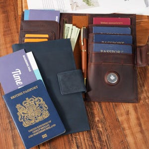 May include: A brown leather travel wallet with multiple compartments, holding passports, boarding passes, and cash. A blue passport and a blue notebook are visible. A gold pen is placed inside the wallet. The wallet is open, displaying its contents.