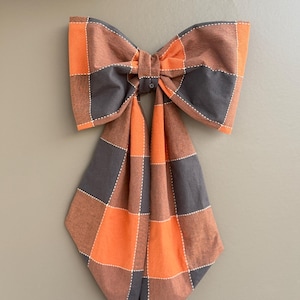 May include: A large, decorative bow made of fabric, featuring a plaid pattern in shades of orange, gray, and brown. The bow is stitched with white thread and hangs against a neutral-colored wall. The bow is a decorative accent.