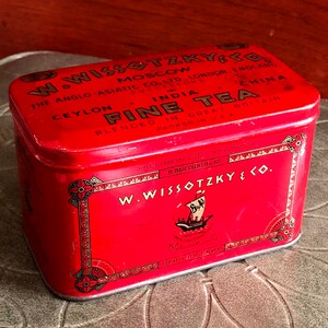 May include: A vintage red rectangular tea tin with the text "W. Wissotzky & Co." and "Fine Tea." The tin has a decorative border and a ship illustration. The text includes locations like Moscow, London, and Ceylon.