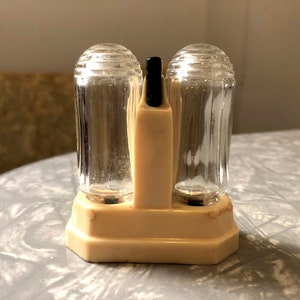 May include: Vintage salt and pepper shaker set. The set features two clear glass shakers with ribbed sides and clear, domed lids. They are held in a cream-colored plastic holder with a black handle. The base is a cream-colored, octagonal shape.