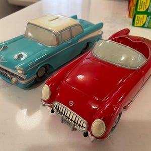 May include: Two vintage car-shaped coin banks. One is a teal-colored classic car with a cream-colored roof. The other is a red convertible with a silver windshield. Both have coin slots on top and are displayed on a white surface.