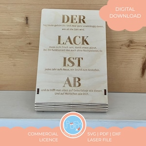 Könnte beinhalten: Helle Holzplakette mit graviertem Text. Die Wörter "DER", "LACK", "IST" und "AB" sind deutlich zu sehen, zusammen mit weiteren Sätzen. Die rechteckige Plakette enthält auch den Schriftzug "DIGITALER DOWNLOAD".
