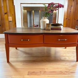 May include: A rectangular coffee table with a dark wood top and a vibrant orange base featuring two drawers. Each drawer is equipped with a horizontal handle that has a dark center and gold-toned ends. The table stands on four tapered legs.
