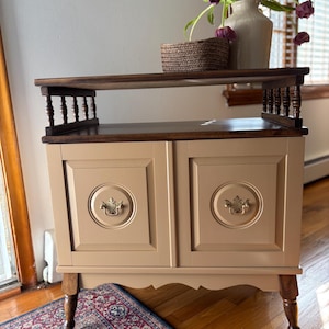 May include: A two-tiered wooden cabinet with a dark brown top and a light tan base. The cabinet has two doors with decorative hardware and sits on four legs. A woven basket and a vase of purple flowers sit on the top shelf. The cabinet is approximately 30 inches tall.