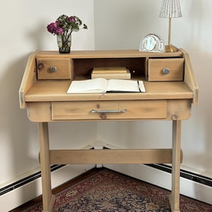 May include: A light brown wooden writing desk featuring a slanted top, one large central drawer with a silver handle, and two smaller upper drawers with round knobs. An open notebook, two books, a glass vase with flowers, and a lamp sit on top.