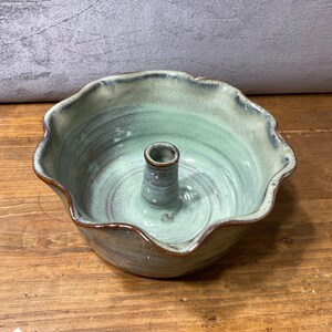 May include: A ceramic candle holder bowl with a scalloped edge and a central pillar. The piece features a light sage green glaze with subtle blue-grey undertones and dark brown accents along the rim and the top of the central candle spike.