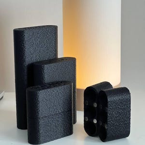 Puede incluir: Una colección de objetos negros, texturizados y rectangulares de varios tamaños. Los objetos están dispuestos sobre una superficie blanca, con una lámpara cilíndrica blanca en el fondo. La lámpara emite una luz cálida y suave.
