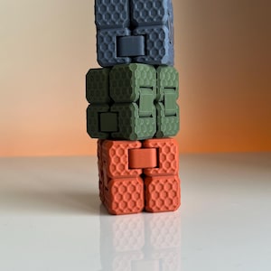 Puede incluir: Una pila de tres cubos fidget en tonos grises, verde oliva y naranja. Cada cubo está compuesto por bloques más pequeños y texturizados con un patrón de panal. Los cubos están conectados por bisagras, lo que permite el movimiento.